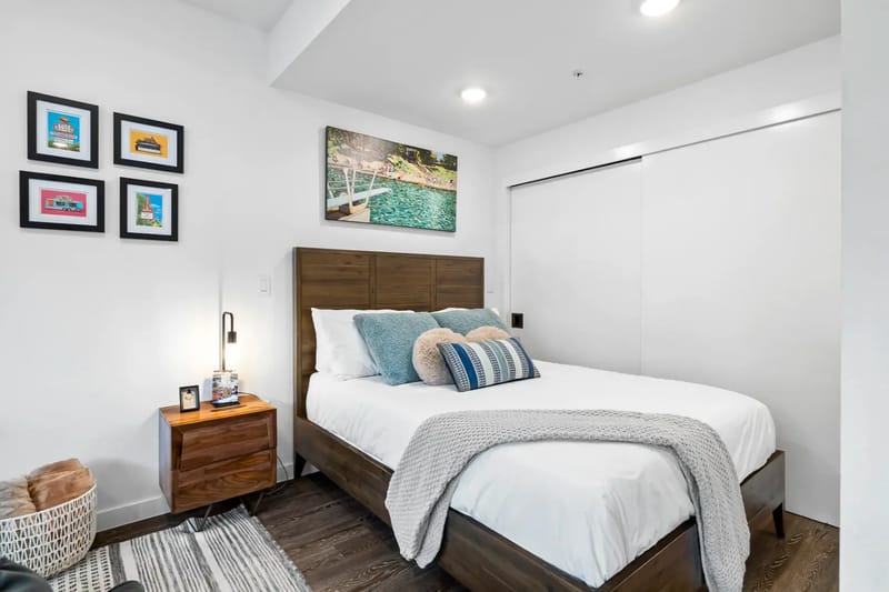 Un dormitorio moderno con cama de madera, ropa de cama blanca, una mesa de noche con lámpara y arte en la pared. Ambiente acogedor con iluminación suave.
