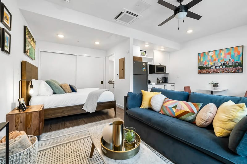 Sala acogedora con sofá azul, cojines amarillos, televisor montado en la pared, lámpara y una ventana desde la que se ve el follaje verde del exterior. Detalles decorativos modernos.