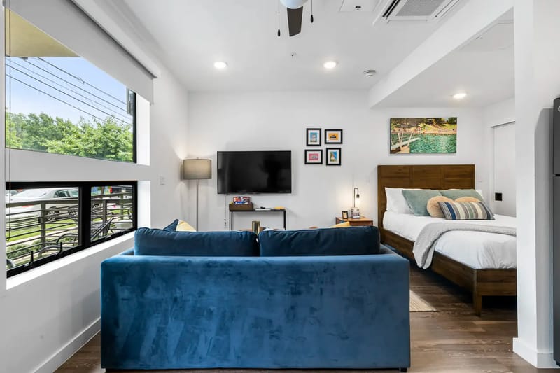 Un apartamento tipo estudio moderno con sofá azul, televisor montado en la pared, cama y cuadros enmarcados. Las ventanas grandes proporcionan luz natural.