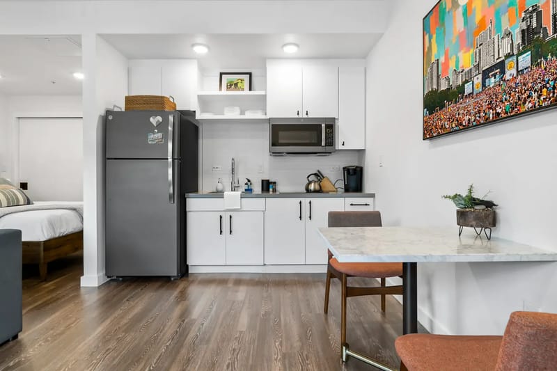 Apartamento tipo estudio moderno con cocina compacta equipada con refrigerador, estufa y microondas. Cerca hay una mesa pequeña con sillas sobre un piso de madera.