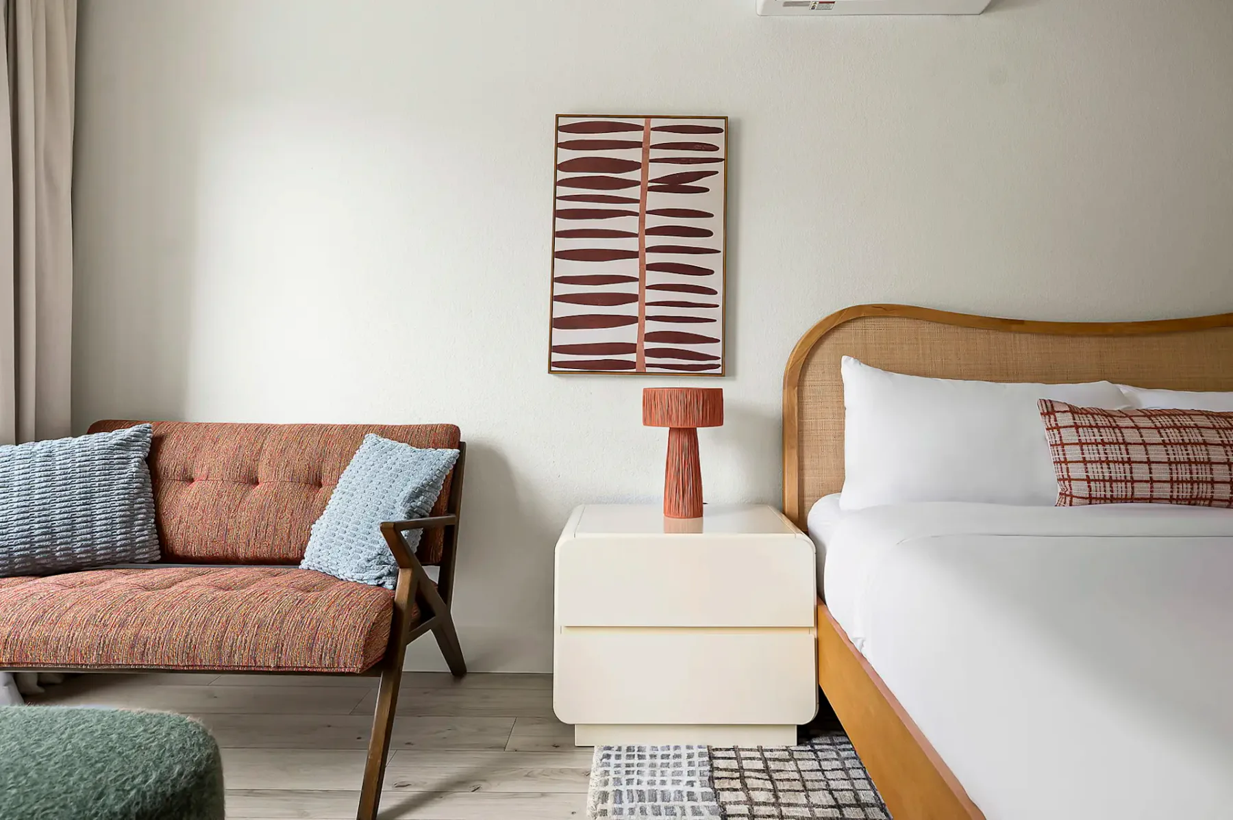 Dormitorio elegante con una cabecera de mimbre, ropa de cama blanca, arte abstracto en la pared, una lámpara moderna y un sofá acogedor adornado con cojines estampados.