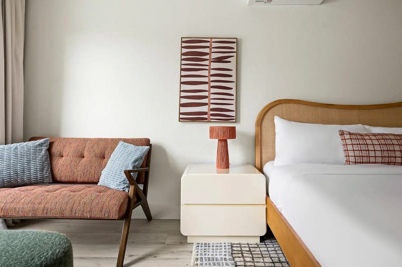 Dormitorio elegante con una cabecera de mimbre, ropa de cama blanca, arte abstracto en la pared, una lámpara moderna y un sofá acogedor adornado con cojines estampados.