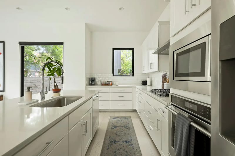 Cocina moderna con gabinetes blancos, electrodomésticos de acero inoxidable, fregadero en la isla y una ventana desde la que se ven árboles verdes.