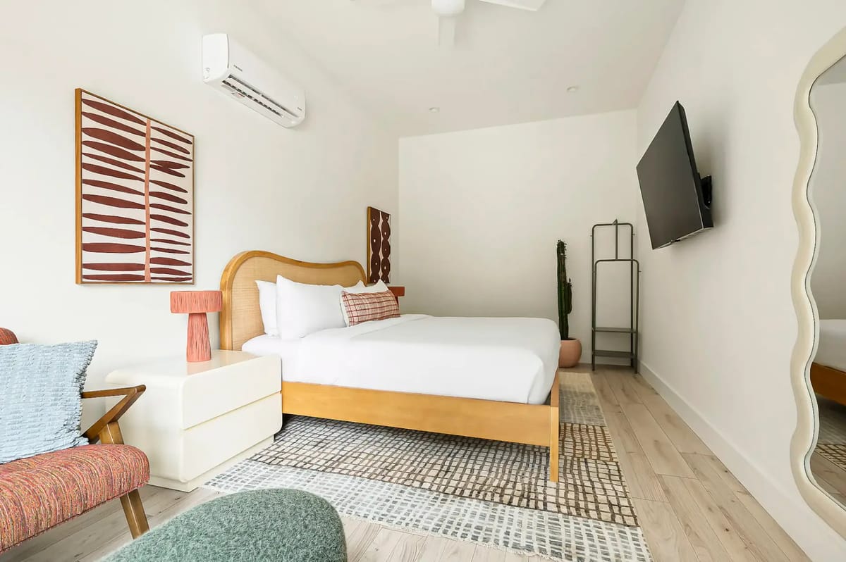 Dormitorio moderno con una cama de madera, ropa de cama blanca, arte en la pared, un espejo grande, un televisor montado en la pared y decoración minimalista sobre un piso de madera clara.