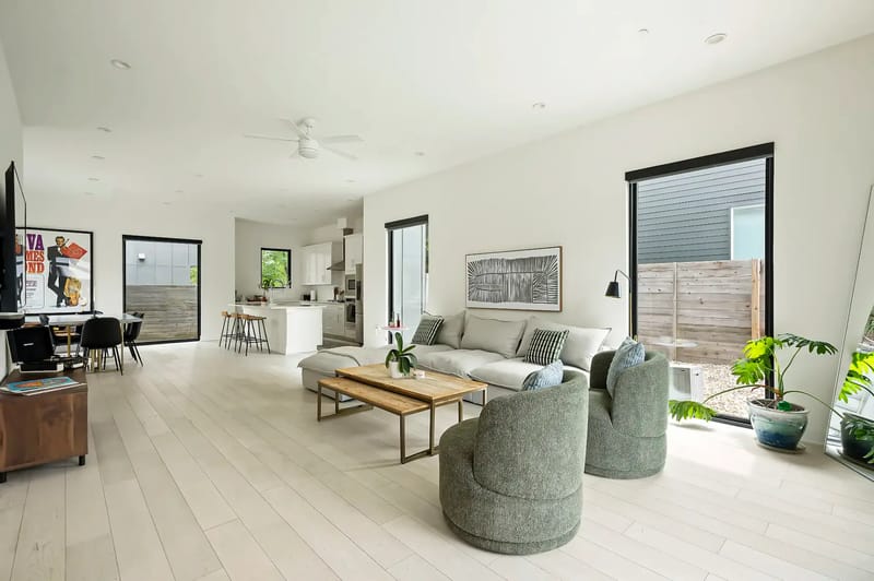 Sala de estar moderna con paredes blancas, piso de madera clara, un sofá gris, sillones verdes, mesa de centro, área de cocina y ventanas grandes.