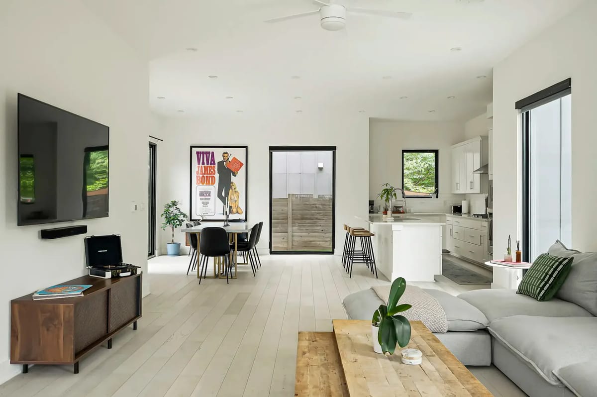 Sala de estar moderna de concepto abierto con cocina, un sofá seccional gris, un televisor montado en la pared, muebles de madera y ventanas grandes que permiten la entrada de luz natural.