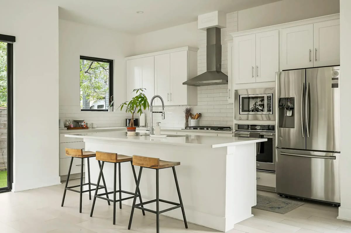 Cocina moderna con gabinetes blancos, electrodomésticos de acero inoxidable y una isla central con tres bancos de madera. Las ventanas grandes proporcionan luz natural.