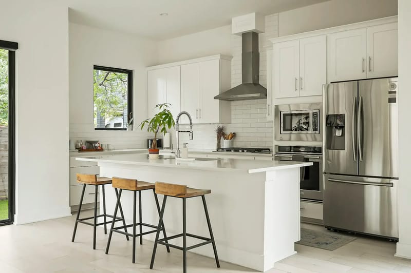 Cocina moderna con gabinetes blancos, electrodomésticos de acero inoxidable y una isla central con tres bancos de madera. Las ventanas grandes proporcionan luz natural.