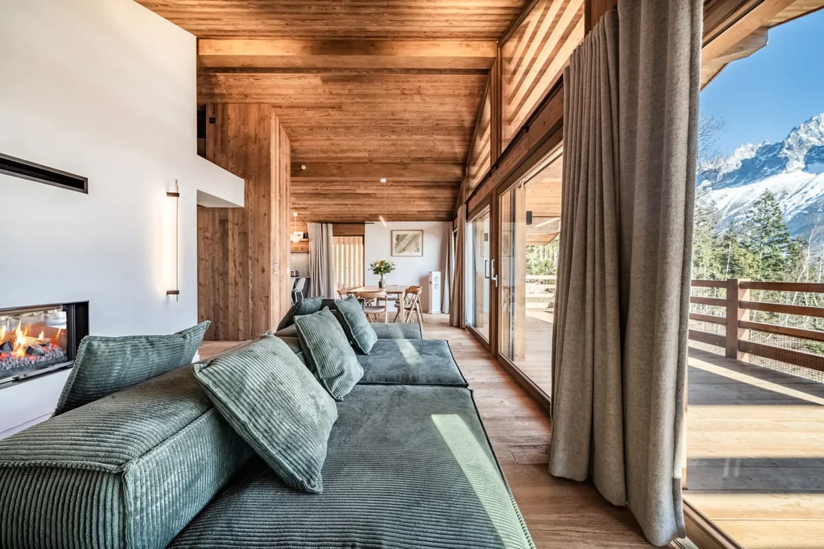 Amplio salón con grandes sofás mullidos, vigas de madera, una chimenea moderna y puertas de cristal que dan a un balcón con vistas a las montañas.