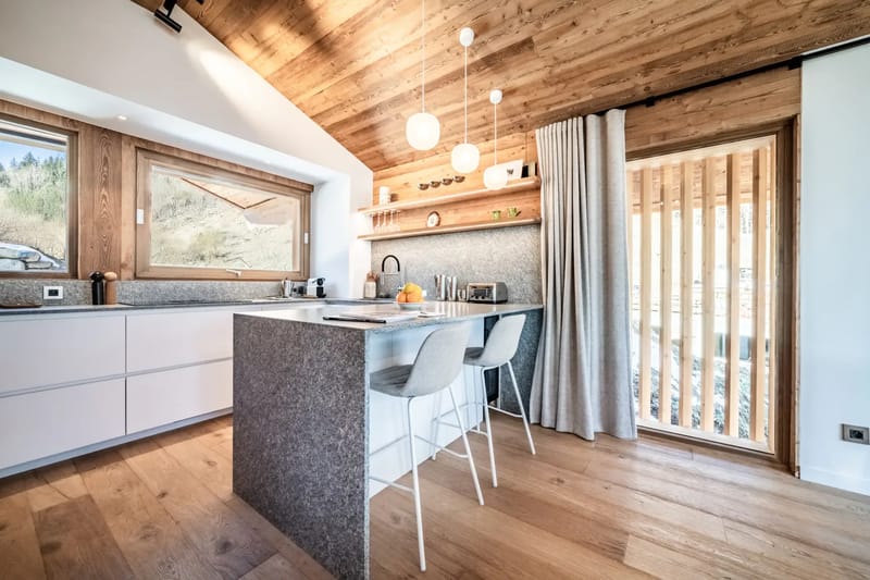 Cocina moderna con techo inclinado de madera, isla central, taburetes y una gran ventana con vistas.