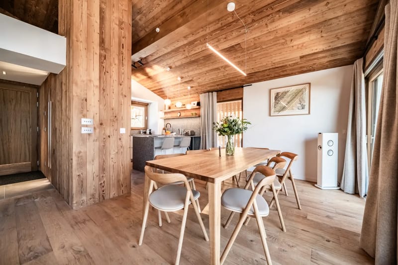 Zona de comedor de madera con mesa y sillas, grandes ventanas y vistas a montañas nevadas.