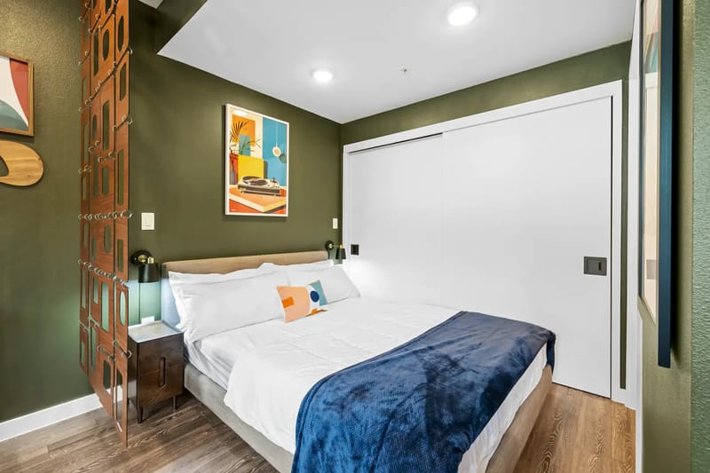 Área de descanso moderna con cama doble, paredes verdes, manta azul, obras de arte coloridas y piso de madera.