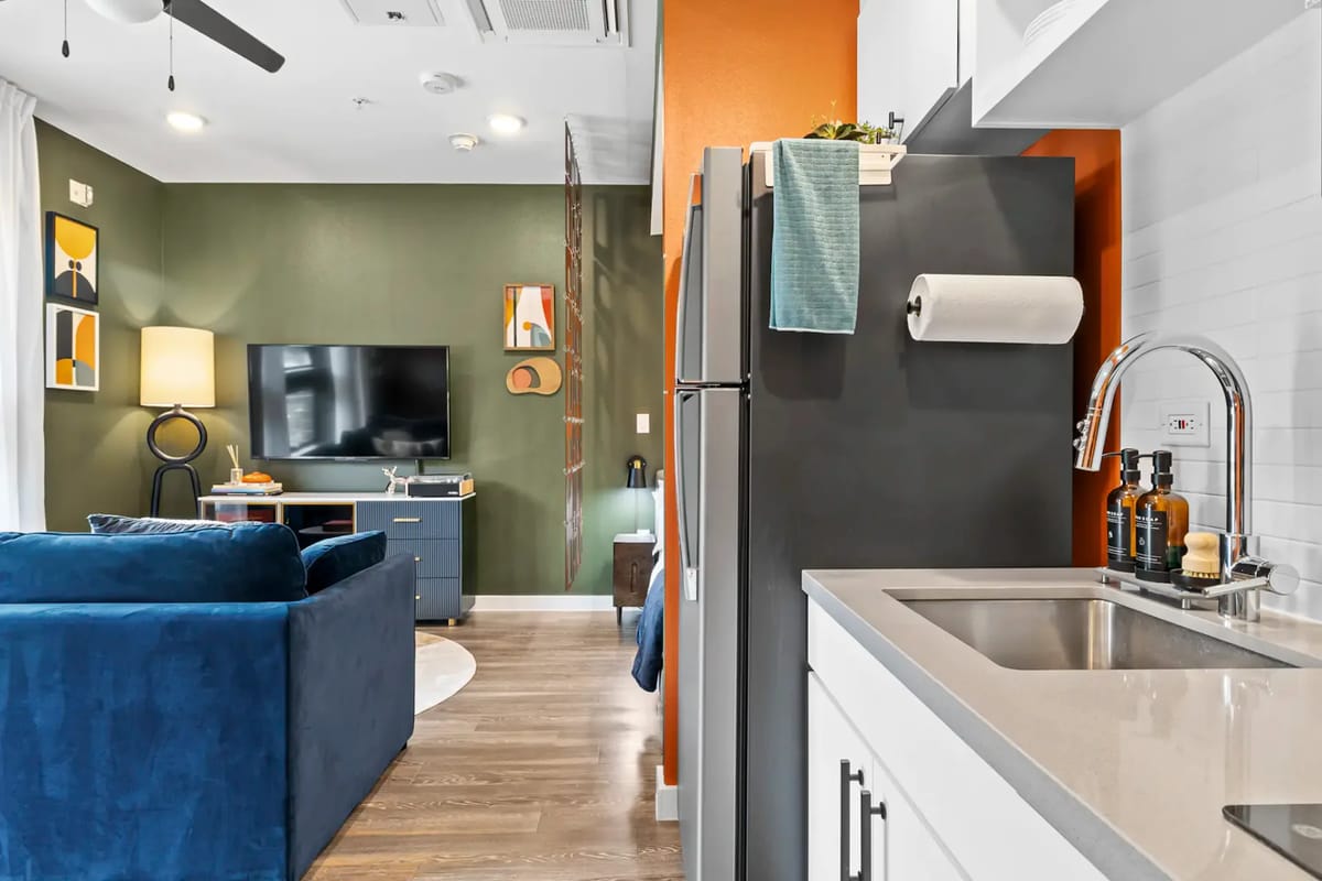 Modern living space with blue sofa, wall-mounted TV, colorful art, and a kitchen area with stainless steel appliances.