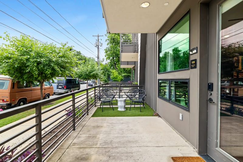 Patio exterior con sillas de metal y una mesa pequeña sobre césped artificial junto al edificio.