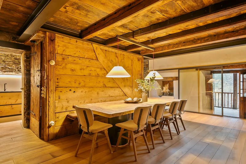 Comedor rústico con una mesa de madera, seis sillas, lámparas colgantes, vigas vistas y acceso al balcón.