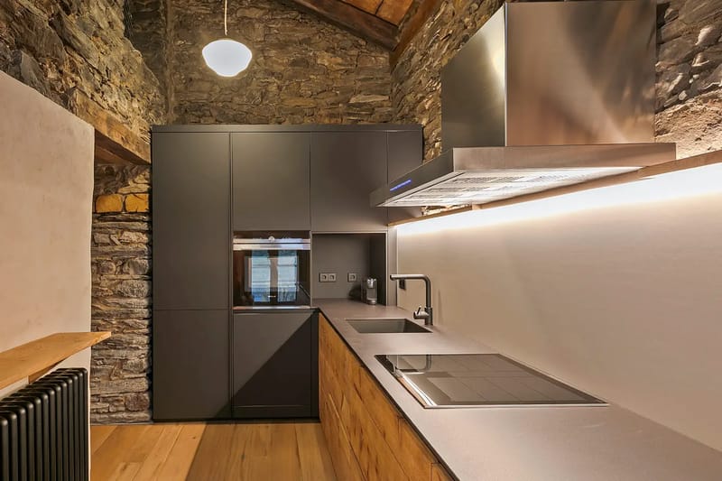 Cocina moderna con paredes de piedra, armarios oscuros, una campana extractora de acero inoxidable y un fregadero y una placa de cocina integrados.