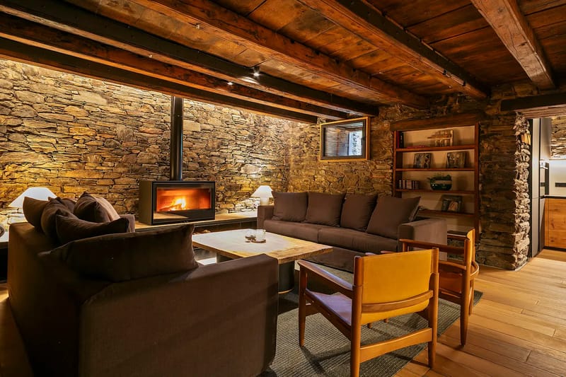 Salón acogedor con una pared decorativa de piedra, sofás oscuros, techo de madera y una chimenea encendida.