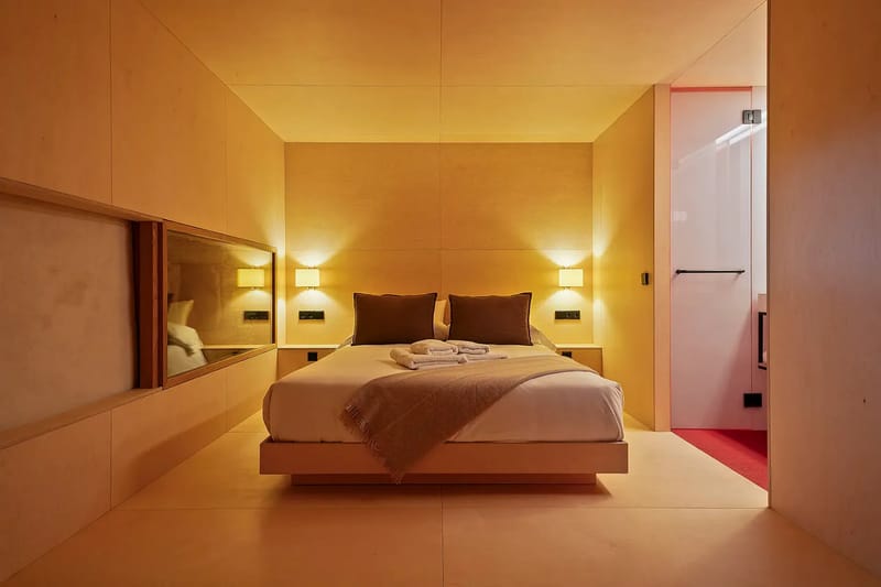 Dormitorio con paredes de madera, una cama doble, iluminación cálida y un baño visible al fondo.