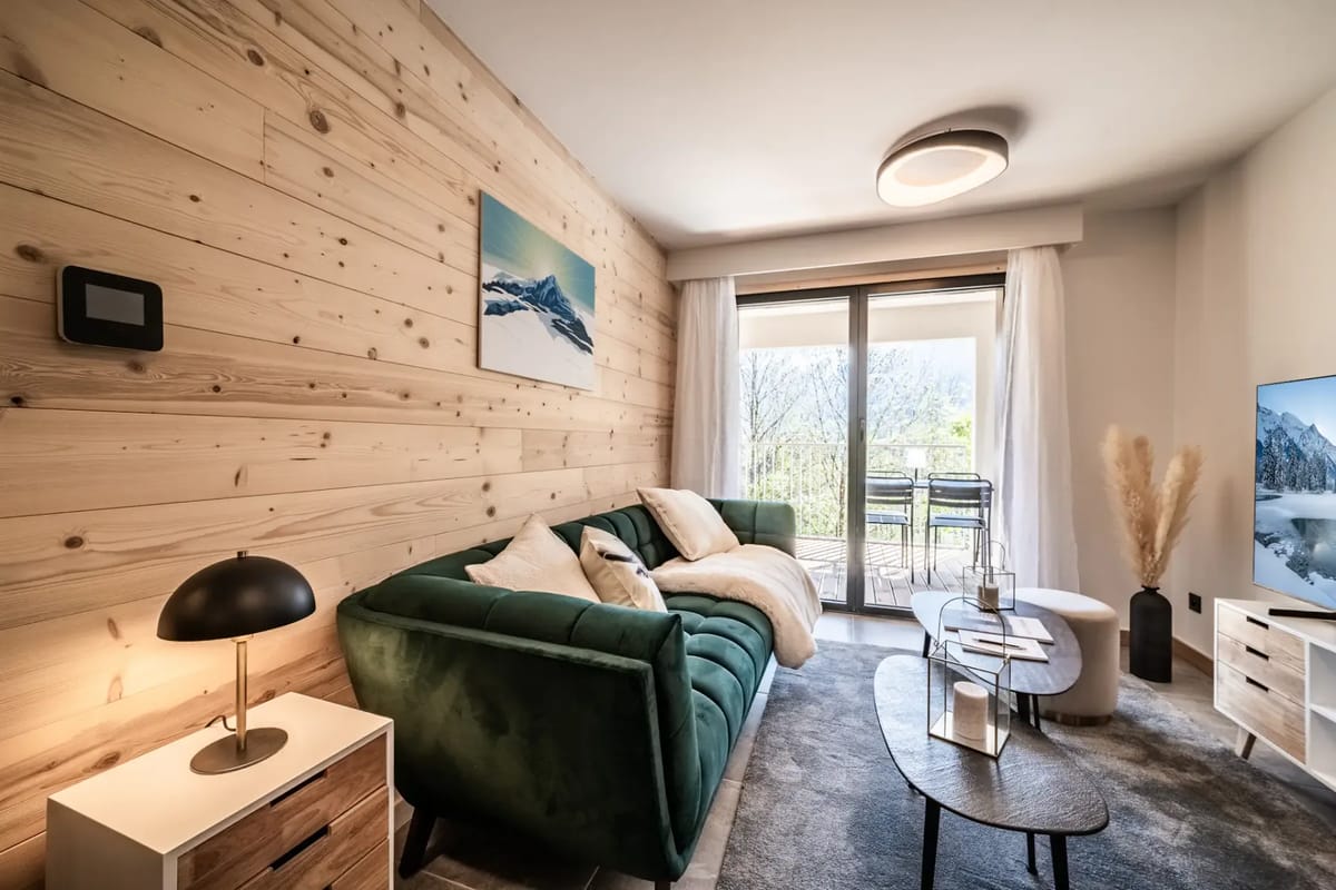 Modern living room with a green sofa, TV displaying a mountain scene, wooden accent wall, coffee table, and sliding glass door leading to a balcony with outdoor seating.