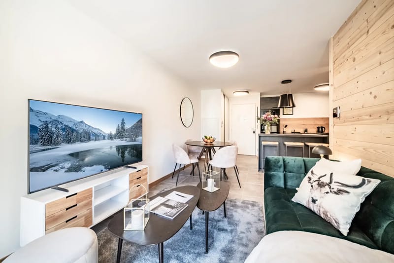 Modern living room with a green sofa, TV displaying a mountain scene, wooden accent wall, coffee table, and sliding glass door leading to a balcony with outdoor seating.