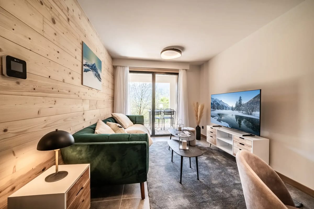 Modern living room with a green sofa, TV displaying a mountain scene, wooden accent wall, coffee table, and sliding glass door leading to a balcony with outdoor seating.