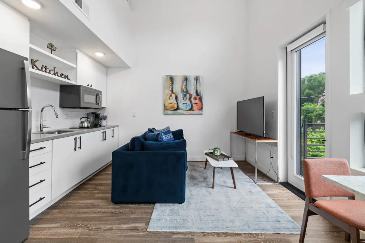 Modern studio apartment with a kitchen, blue sofa, TV, artwork of guitars, and large windows overlooking greenery. Light wood flooring.