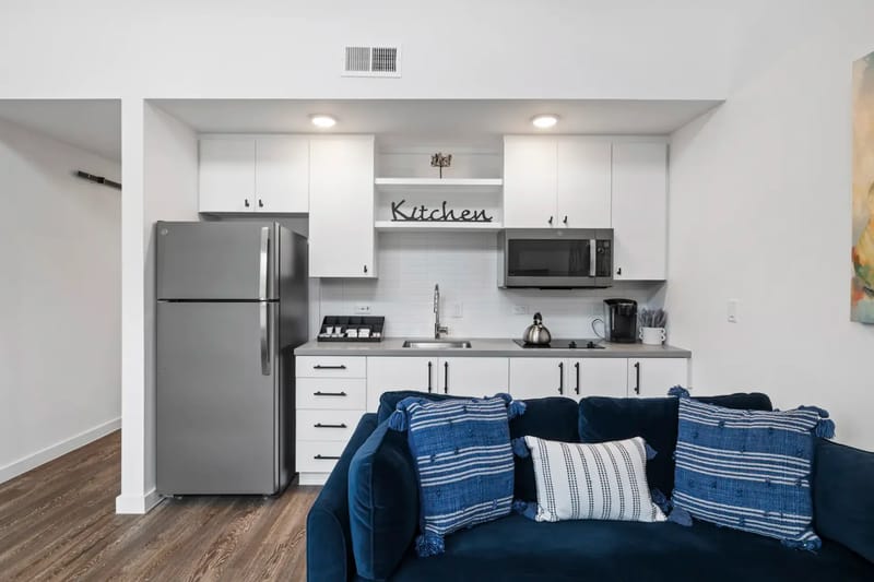 Cocina moderna compacta con gabinetes blancos, electrodomésticos de acero inoxidable y un sofá azul con cojines estampados en primer plano.