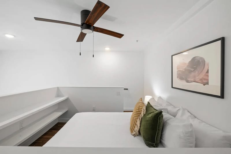 Dormitorio minimalista con una cama blanca, almohadas verdes, arte en la pared, ventilador de techo y estantes empotrados en un espacio luminoso y aireado.
