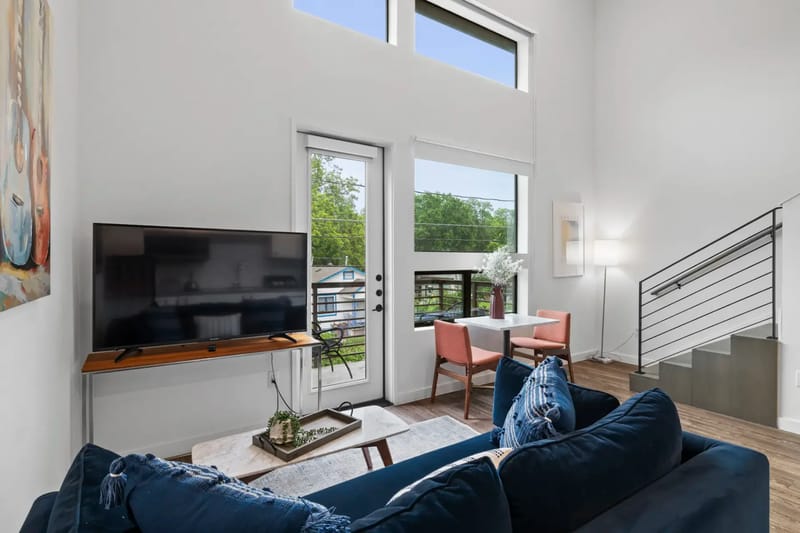Sala de estar moderna con un sofá azul marino, televisor, mesa de comedor pequeña, lámpara de pie y escalera. Las ventanas grandes proporcionan luz natural y vistas al exterior.