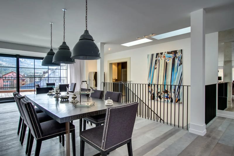 Modern living room with high ceilings, large windows overlooking a snowy landscape, dark green sofas, a wooden coffee table, and artwork of a snowy mountain.