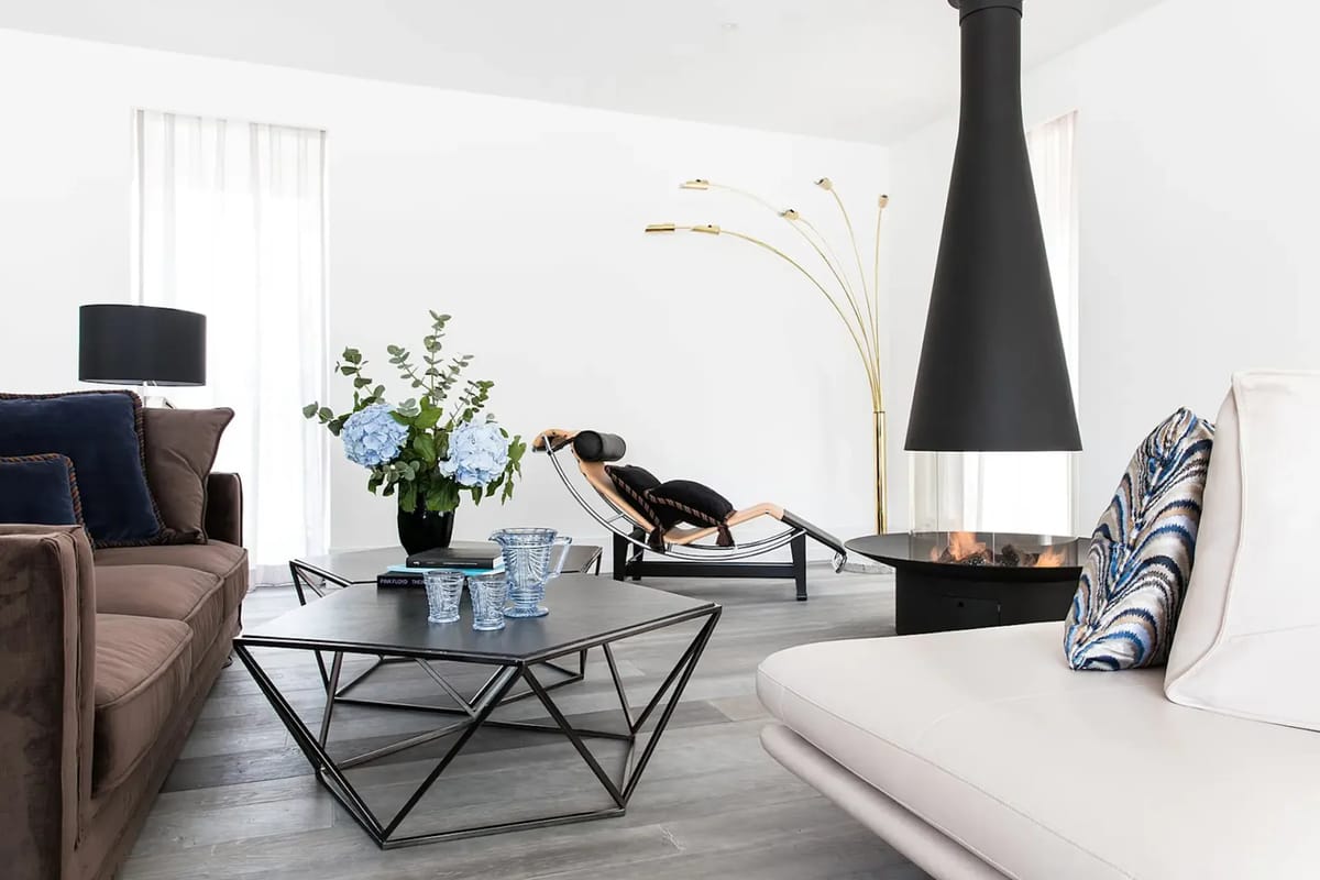 Salón moderno con techos altos, grandes ventanales con vistas a un paisaje nevado, sofás de color verde oscuro, una mesa de centro de madera y una obra de arte que representa una montaña nevada.