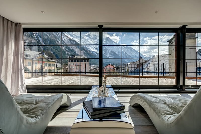 Modern living room with high ceilings, large windows overlooking a snowy landscape, dark green sofas, a wooden coffee table, and artwork of a snowy mountain.
