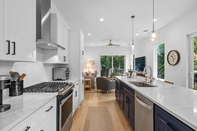 Cocina moderna con gabinetes blancos, electrodomésticos de acero inoxidable, una isla larga con base azul, bancos de bar y pisos de madera que conducen a un patio.