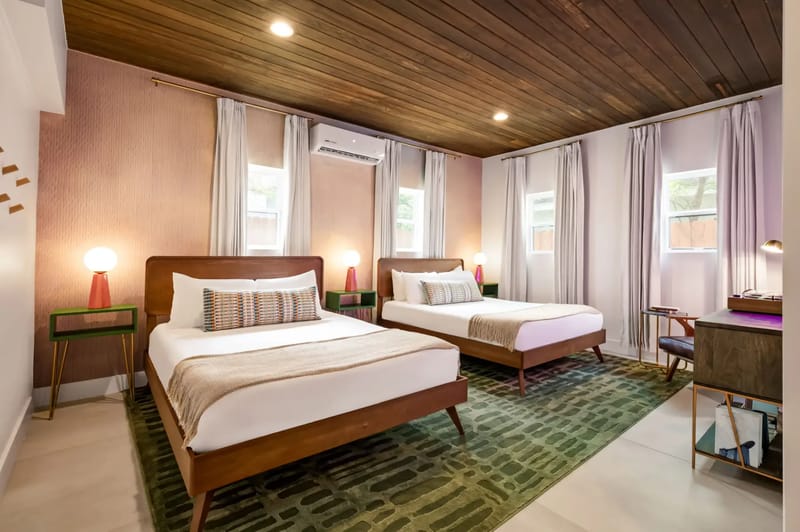 Modern bedroom with two queen beds, wooden ceiling, carpet, and soft lighting. Features two windows and a desk with a chair.