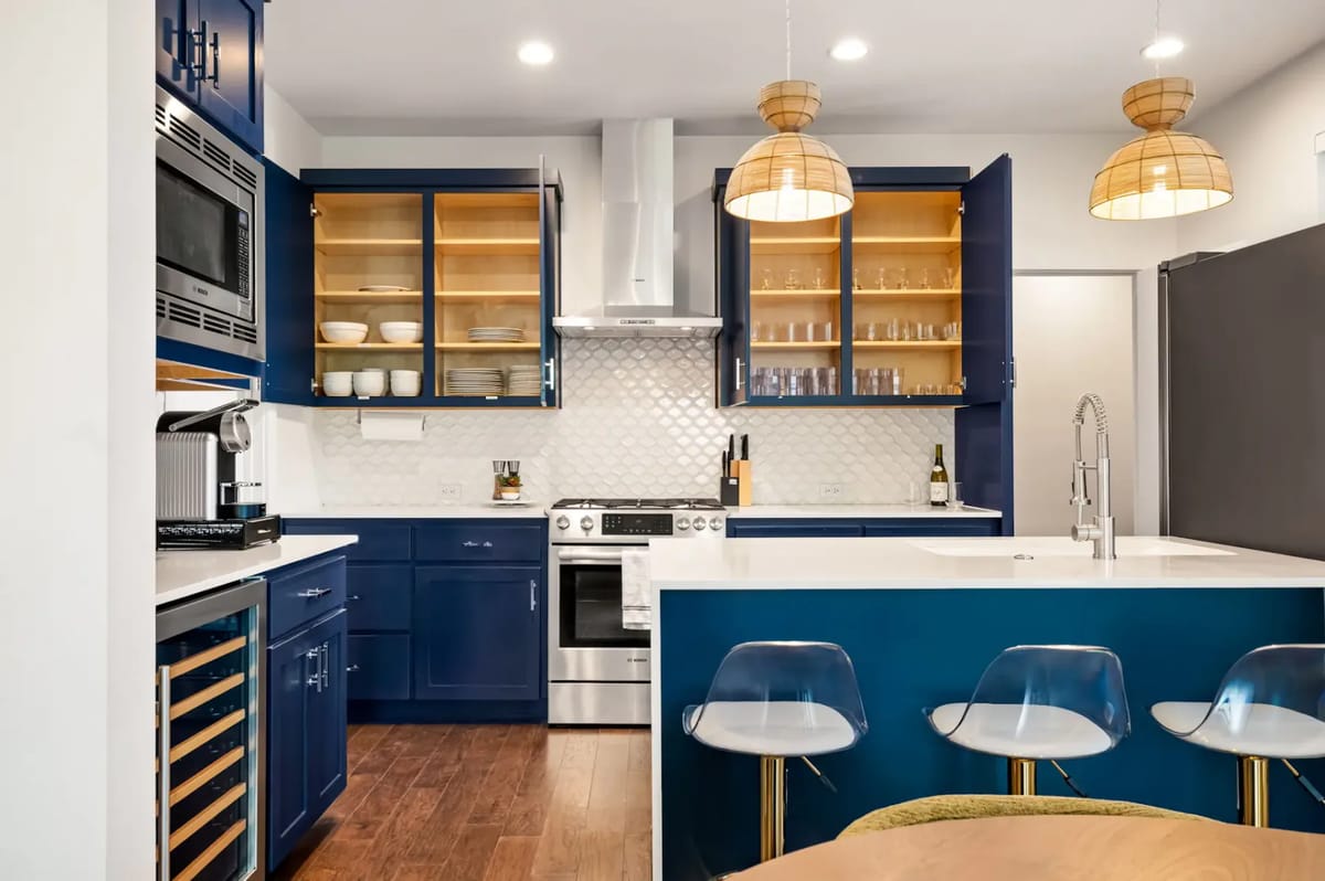 Cocina moderna con gabinetes azul marino, encimeras blancas, estantes abiertos, electrodomésticos de acero inoxidable y lámparas colgantes de mimbre sobre una isla.