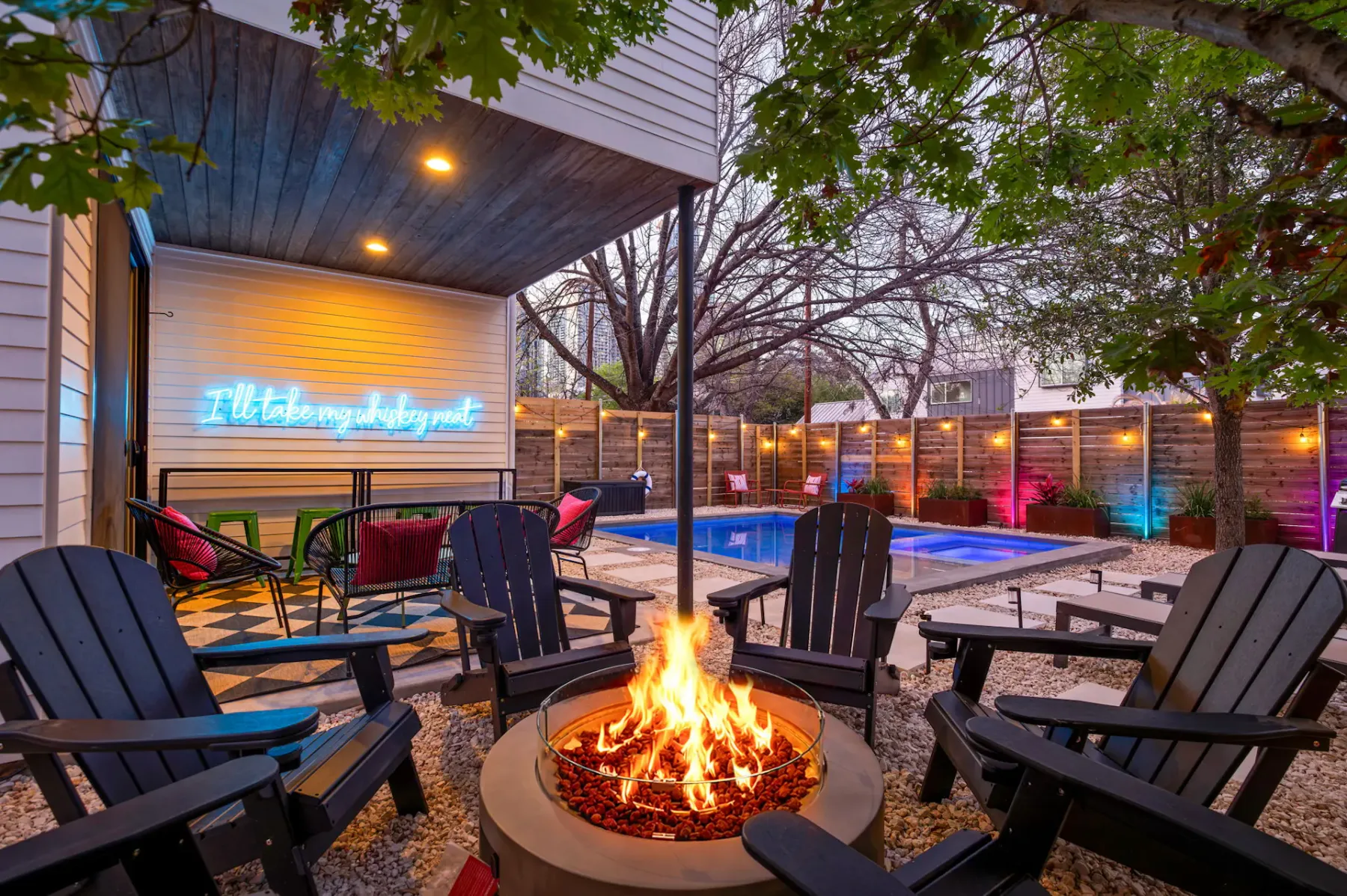 Patio trasero con una fogata rodeada de sillas Adirondack, un letrero de neón en la pared y una piscina iluminada al fondo bajo los árboles.