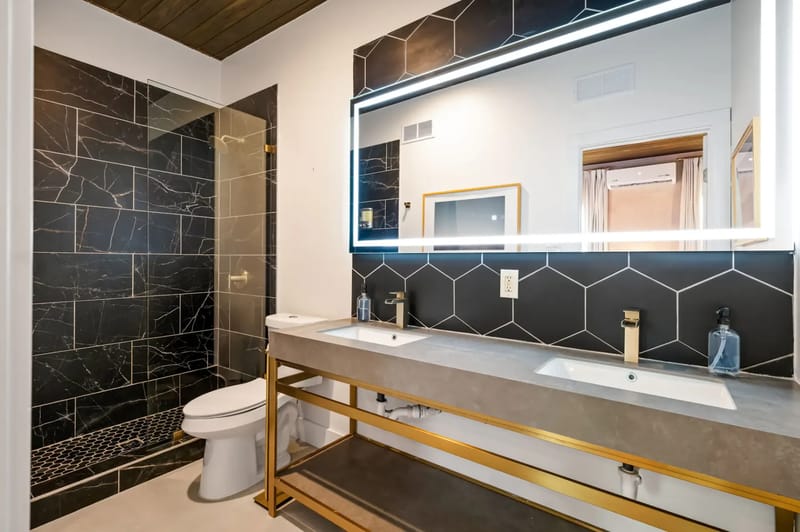 Modern bathroom with a large illuminated mirror, hexagonal black tile wall, dual sinks with gold fixtures, and a black-tiled shower.
