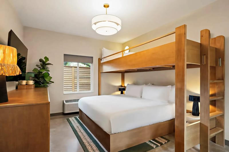 Modern bedroom with a king bed beneath a wooden bunk bed, sleek furniture, a flat-screen TV, and a large window with blinds.