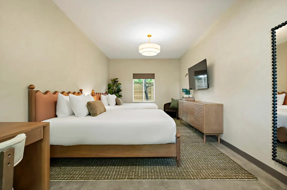 Two double beds, a large mirror, a wall-mounted TV, a wooden dresser, and a window with natural light.