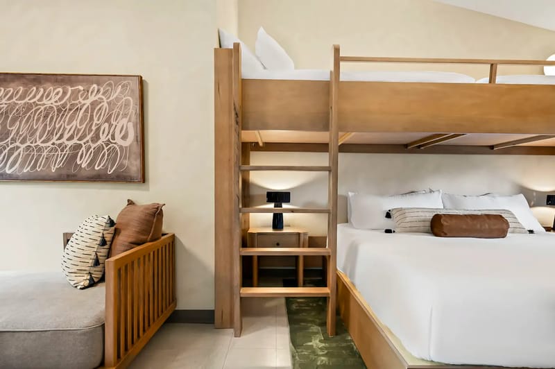 Modern bedroom with a bunk bed, lower queen bed, wooden ladder, wall art, and a cozy seating area with cushions.