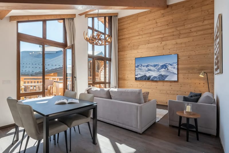 Salón moderno con paredes de madera, grandes ventanas, sofás de color gris claro, una mesa de comedor y un televisor de pantalla plana que muestra una escena de montaña. La luz natural llena el espacio.