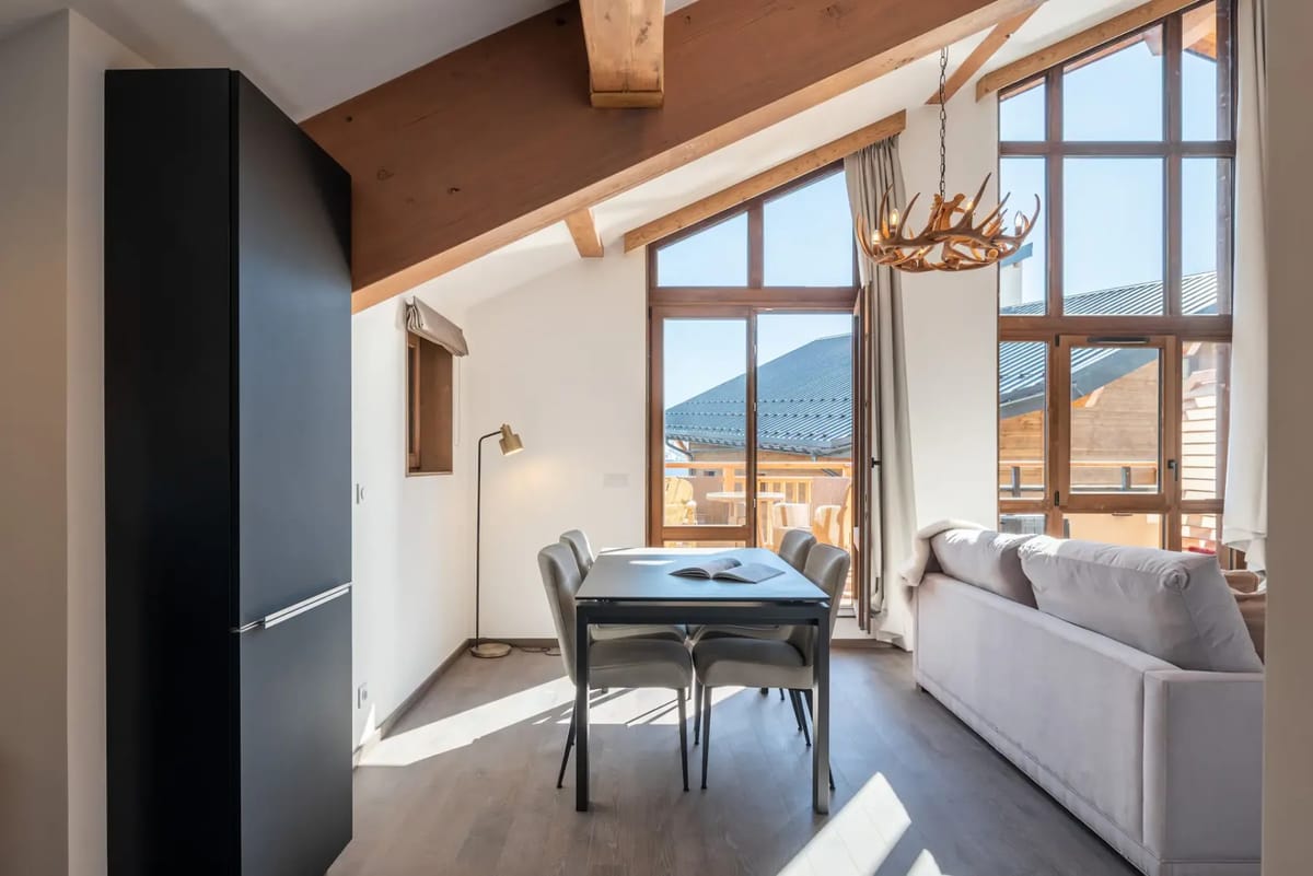 Modern kitchen and dining area with black cabinets, wooden beams, a small window, dining table with four chairs, and an open book on the table.