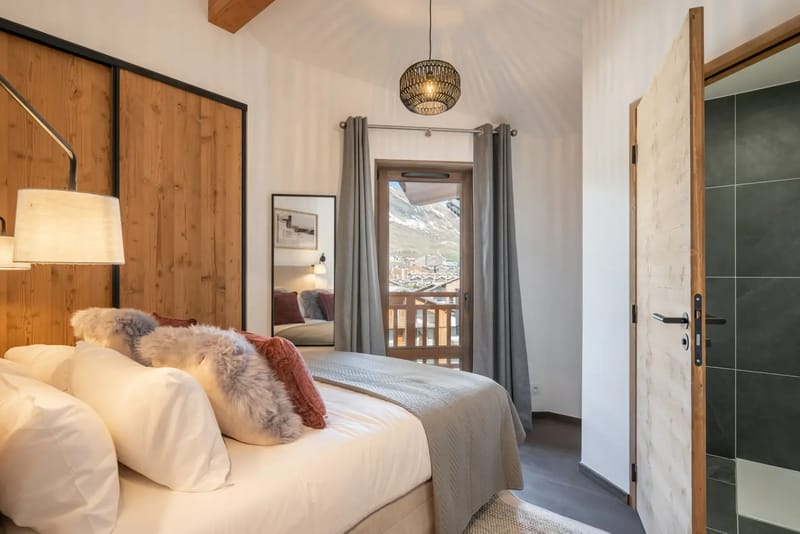 Dormitorio acogedor con una cama bien hecha, almohadas mullidas, detalles de madera y una puerta al balcón que deja ver las montañas; el baño es visible a la derecha.