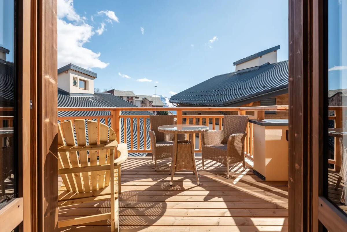 A wooden balcony with outdoor seating, including a round table and chairs, overlooking rooftops under a clear sky.