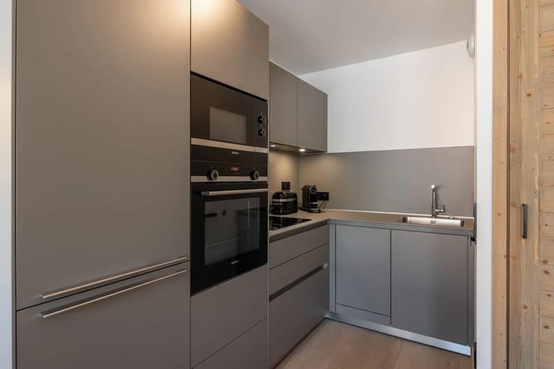 Cocina moderna y compacta con armarios grises, horno empotrado, placa de cocina, microondas y fregadero. Diseño minimalista con líneas limpias y tonos neutros.