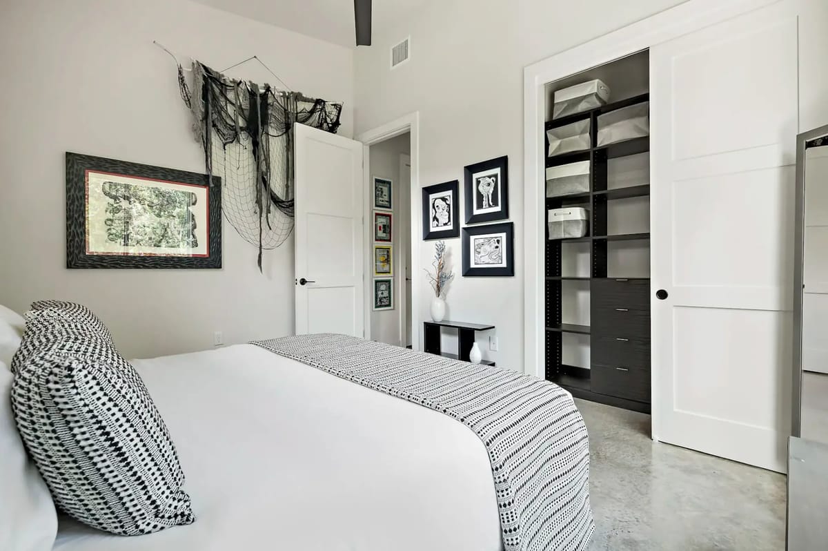Modern bedroom with a white bed, patterned pillows, open closet, framed art, and decorative wall fabric. Neutral tones and minimalist decor.