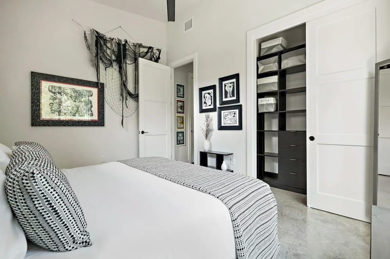 Modern bedroom with a white bed, patterned pillows, open closet, framed art, and decorative wall fabric. Neutral tones and minimalist decor.