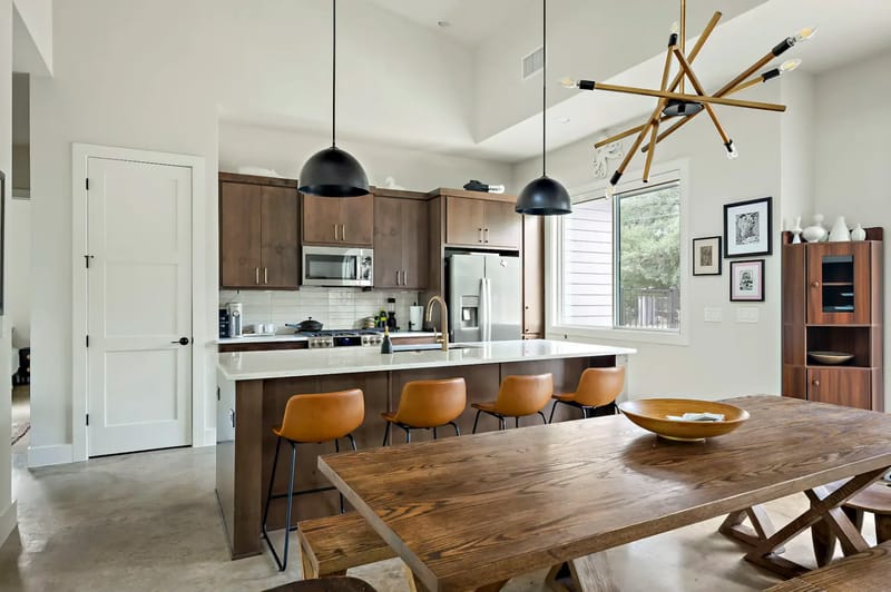 Modern kitchen with wooden cabinets, stainless steel appliances, and a wooden dining table. Three black pendant lights and a unique chandelier hang above.