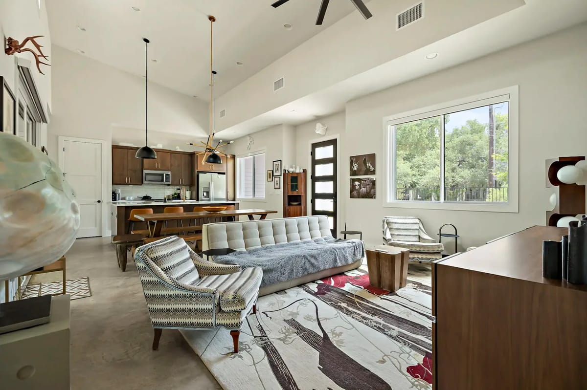 Modern living room with striped chairs, a gray sofa, vibrant rug, and large window. Open kitchen in the background with wooden cabinetry.