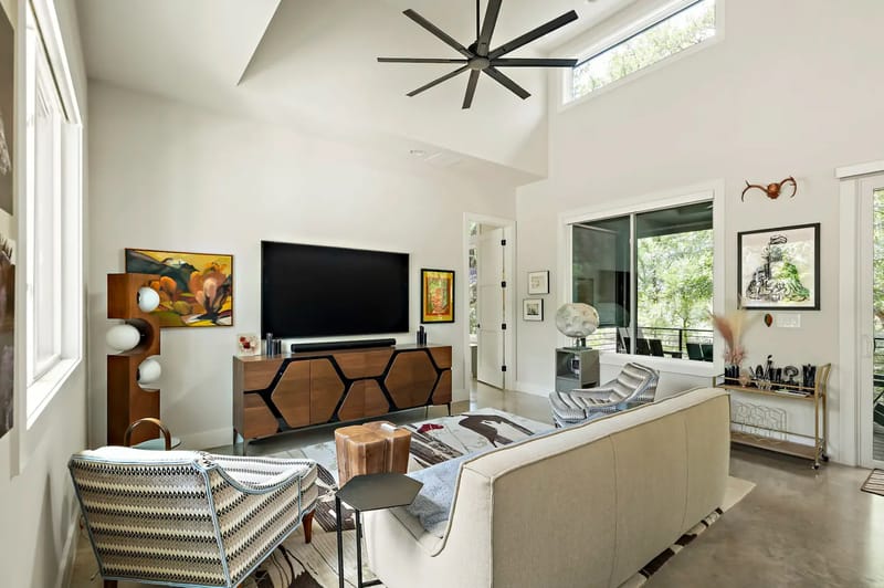 Modern living room with a large TV, patterned chairs, a beige sofa, abstract wall art, decorative items, and a ceiling fan under skylights.